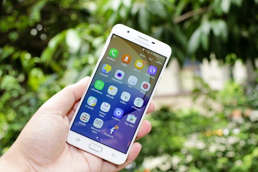 Close-up of a hand holding a smartphone displaying various app icons against a blurred outdoor background.
